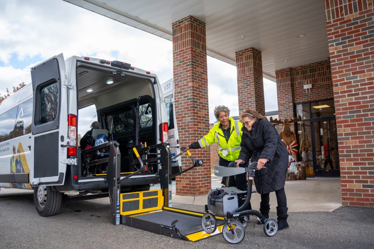 Bus Driver Angie helping participant Annette, who is using a walker, onto a platform to get inside the transportation bus