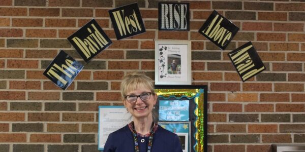 Barb L. smiling in front of a banner and holding a poster board that honors her as a recipient of the David West RISE Above Award