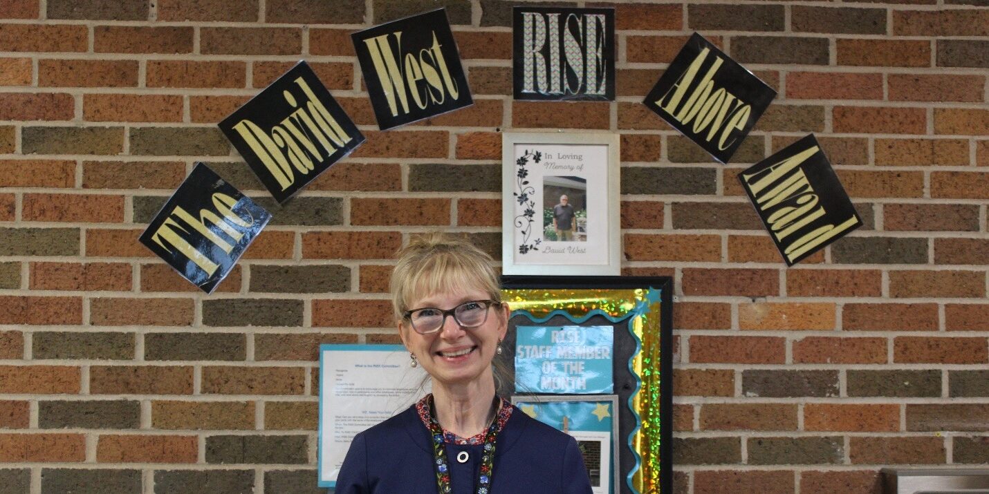 Barb L. smiling in front of a banner and holding a poster board that honors her as a recipient of the David West RISE Above Award