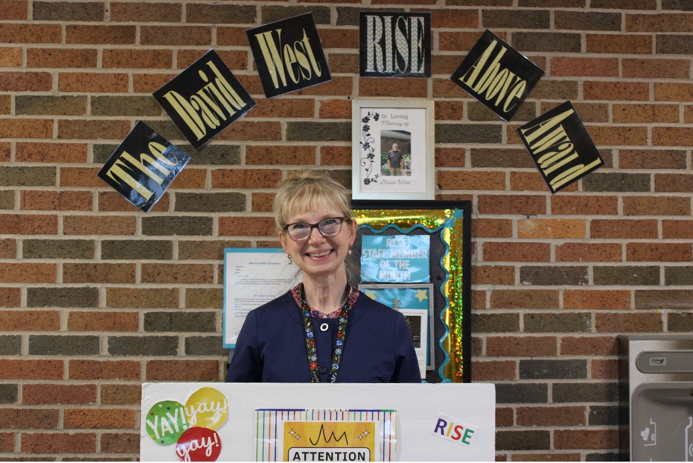 Barb L. smiling in front of a banner and holding a poster board that honors her as a recipient of the David West RISE Above Award