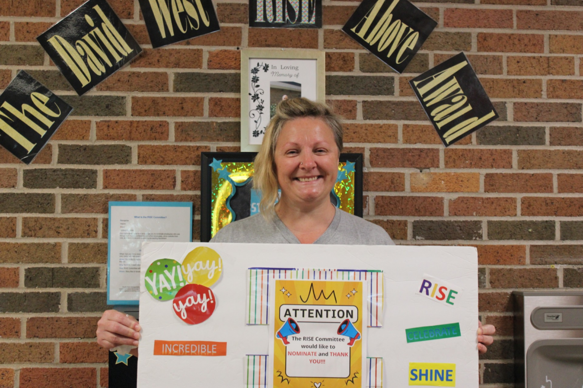 Care Resources June RISE recipient Jamie T. holds a celebratory post board and stands in front of a banner that declares her a recipient of The David West RISE Above Award
