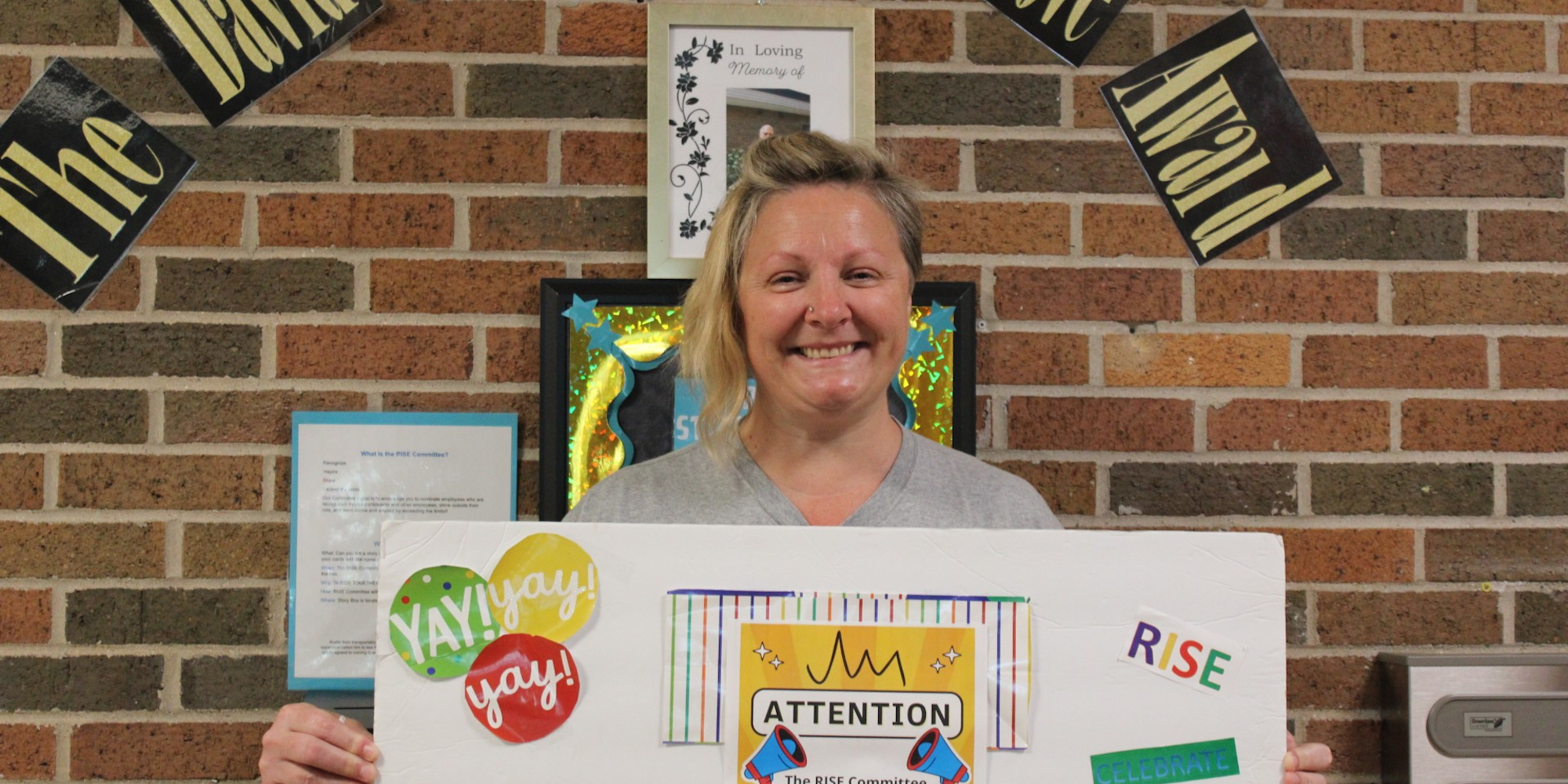 Care Resources June RISE recipient Jamie T. holds a celebratory post board and stands in front of a banner that declares her a recipient of The David West RISE Above Award